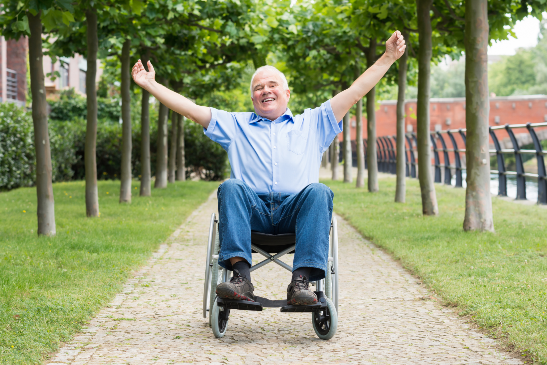 A 40 to 50 year old man sitting a wheelchair with his arms up in joy. The man is outside on a paved path with a line of trees on either side of him.